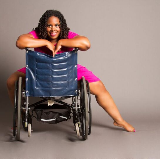 Photo of India Harville, who is smiling into the camera while striking a pose on her backwards-facing wheelchair.